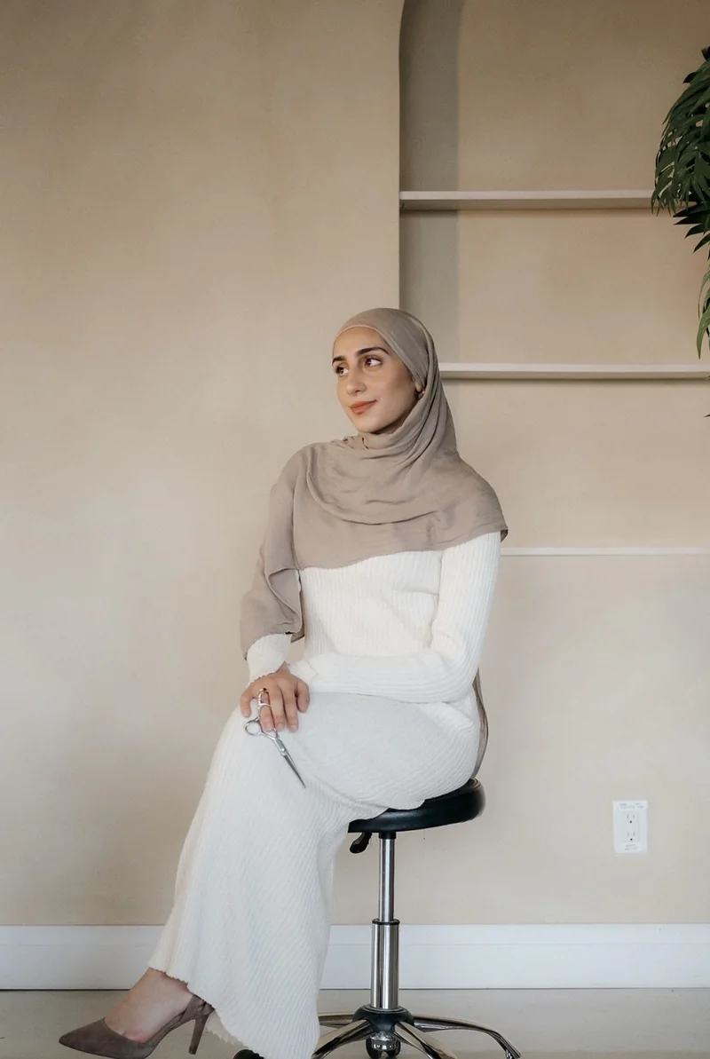 Marwa seated on a styling stool, holding scissors and wearing a taupe hijab and cream outfit.